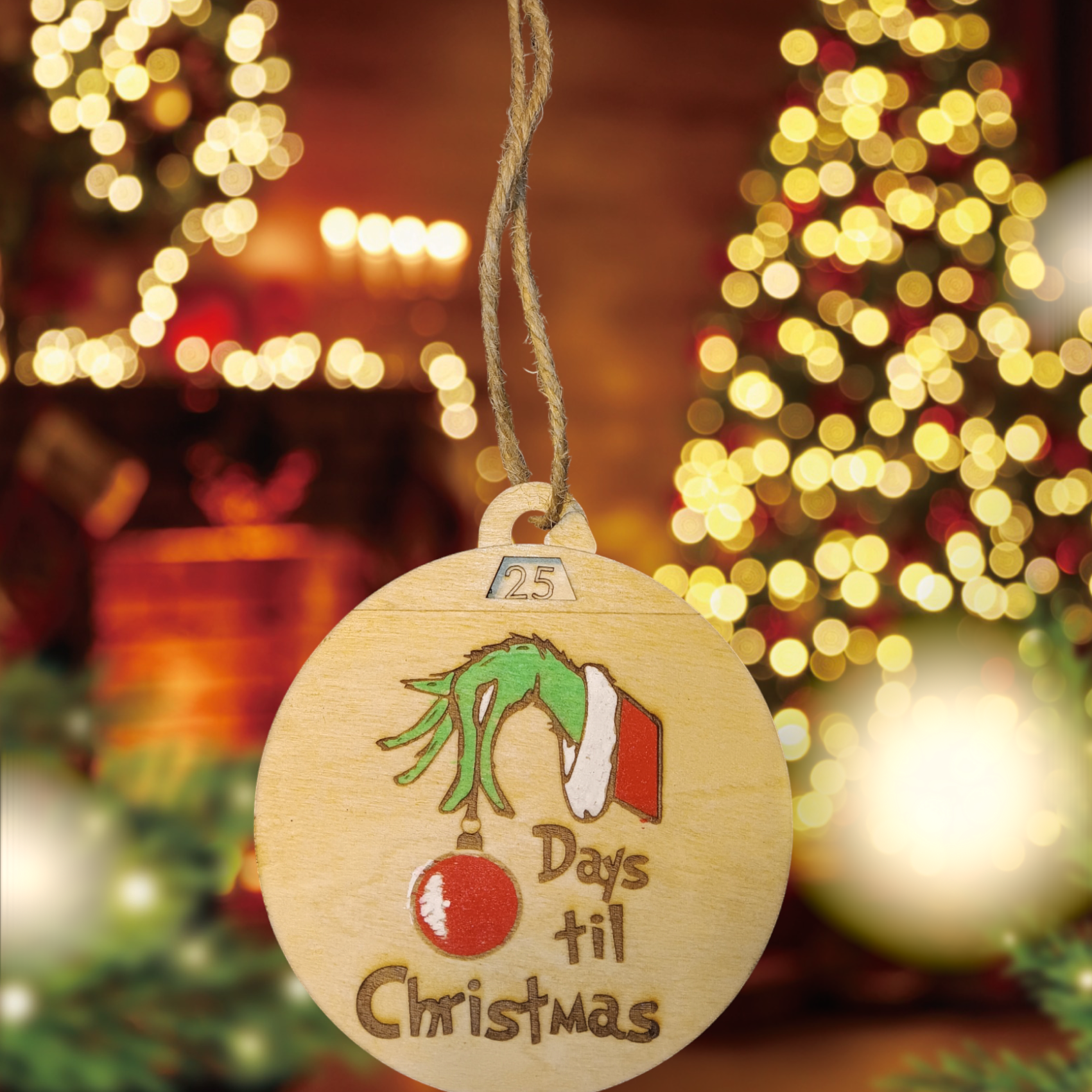 Wooden Christmas ornament featuring a green hand holding a red ornament with the text "Days til Christmas" in front of holiday lights.