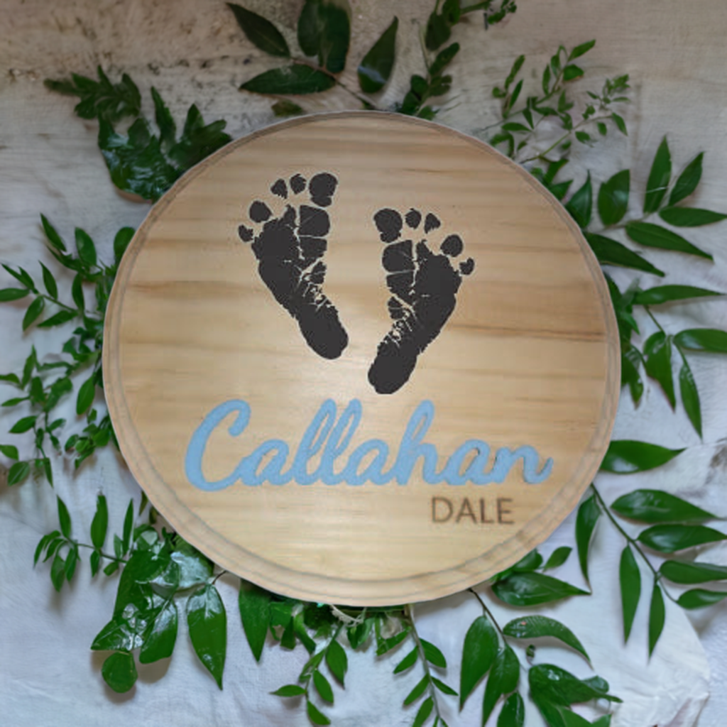 Wooden Baby Footprint Plaque with two black footprints and the name "Callahan Dale," surrounded by green leaves.