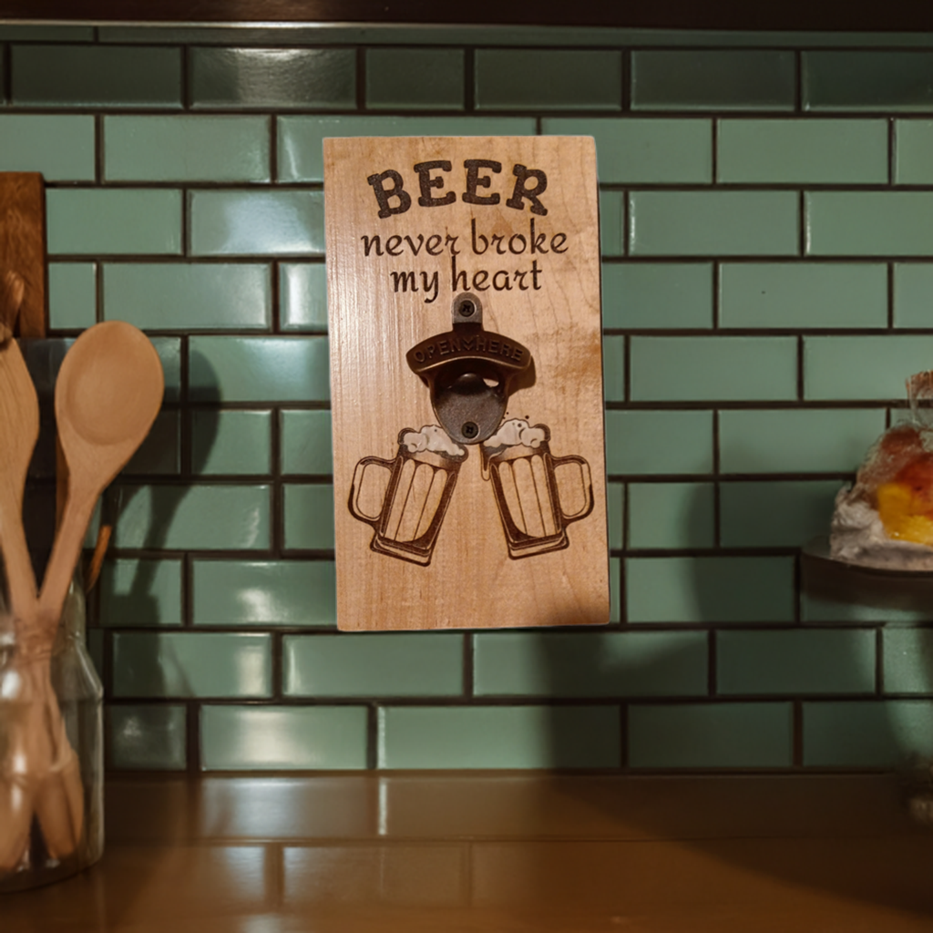 Wooden wall-mounted bottle opener with "Beer never broke my heart" text and engraved beer mugs, displayed on green tile backdrop.