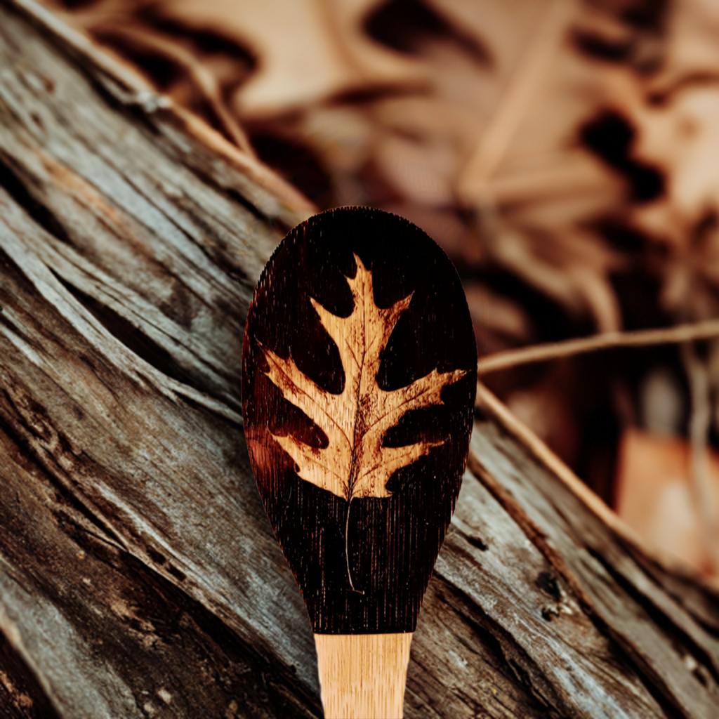 Handmade wood spoon with a carved oak leaf design on the handle, displayed against a rustic wooden background.