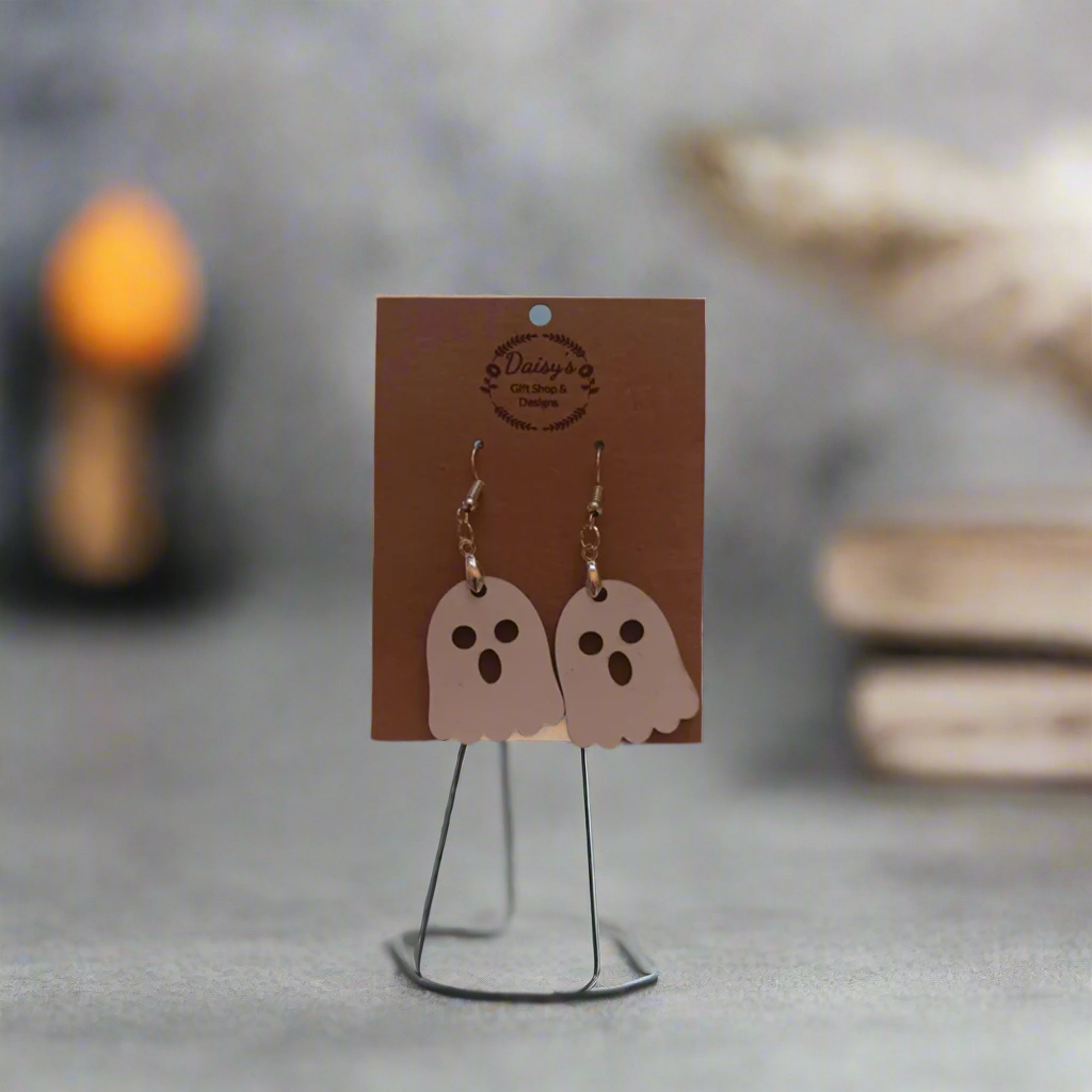 Spooky Earrings featuring white ghost charms hanging on a display stand, blurred background with candles and books.