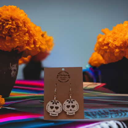 Spooky Earrings product image: Pair of white skull-shaped earrings with heart eyes on a brown card, surrounded by bright orange marigolds.