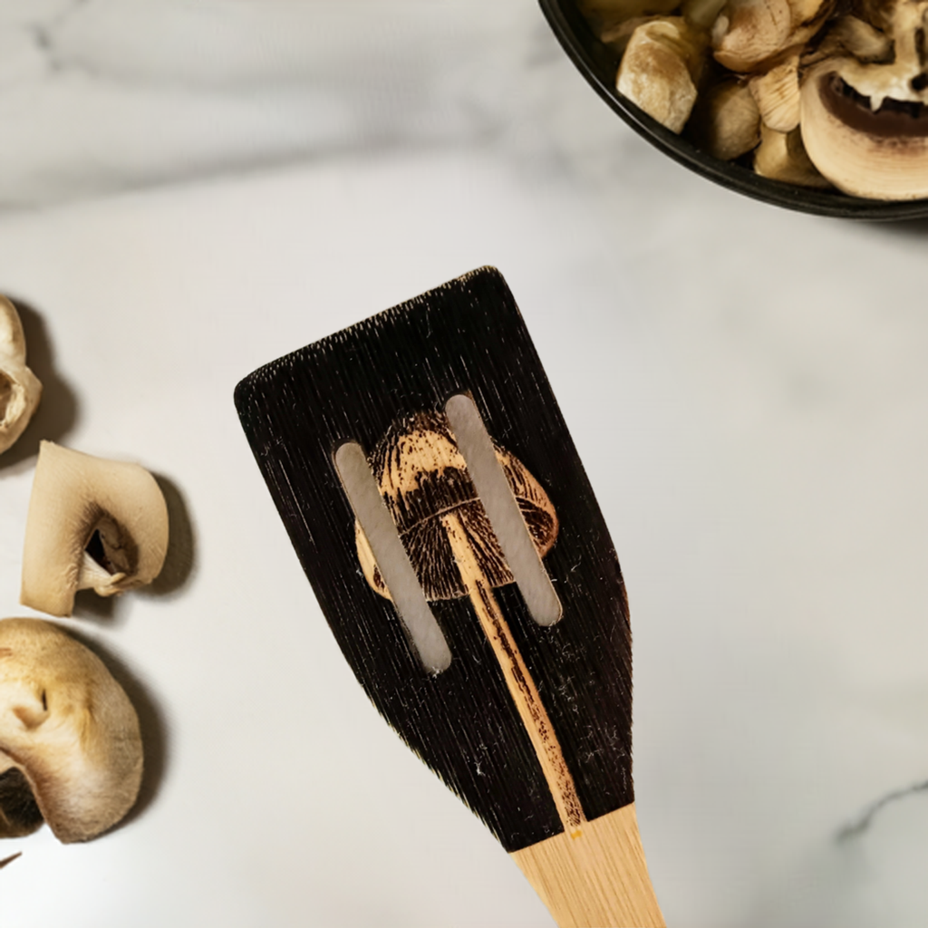 Handmade wood spoon with mushroom artwork on a marble surface, surrounded by sliced mushrooms and a bowl of whole mushrooms.