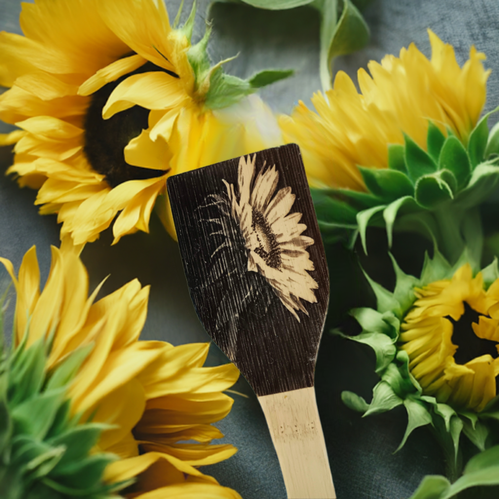 Handmade wooden spoon with sunflower engraving displayed among fresh sunflowers on a gray surface.