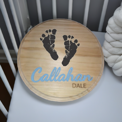 Round wooden plaque with two black baby footprints and the name "Callahan Dale" engraved below in blue text, displayed on a white surface.